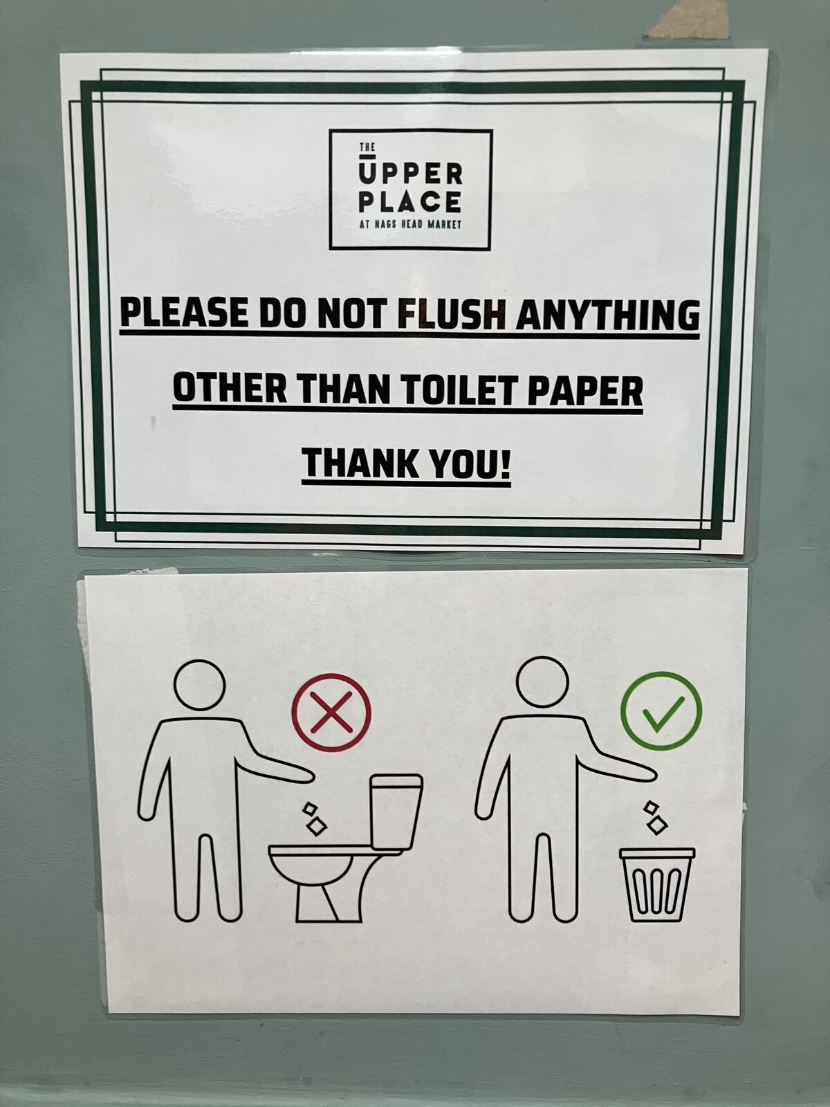 Two signs on a pale green wall: the upper sign says not to flush anything other than toilet paper, and the lower sign shows a pictogram placing waste in a bin rather than a toilet.