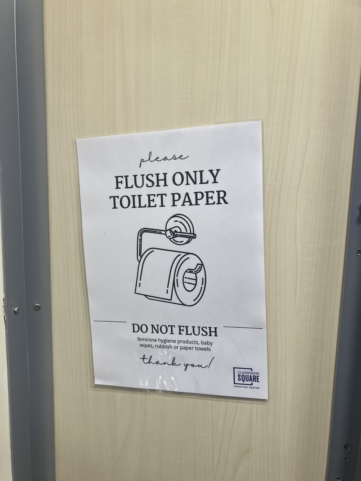 A printed sign on a light wood door with an illustration of a toilet roll, asking users to flush only toilet paper and not feminine hygiene products, baby wipes, rubbish, or paper towels.