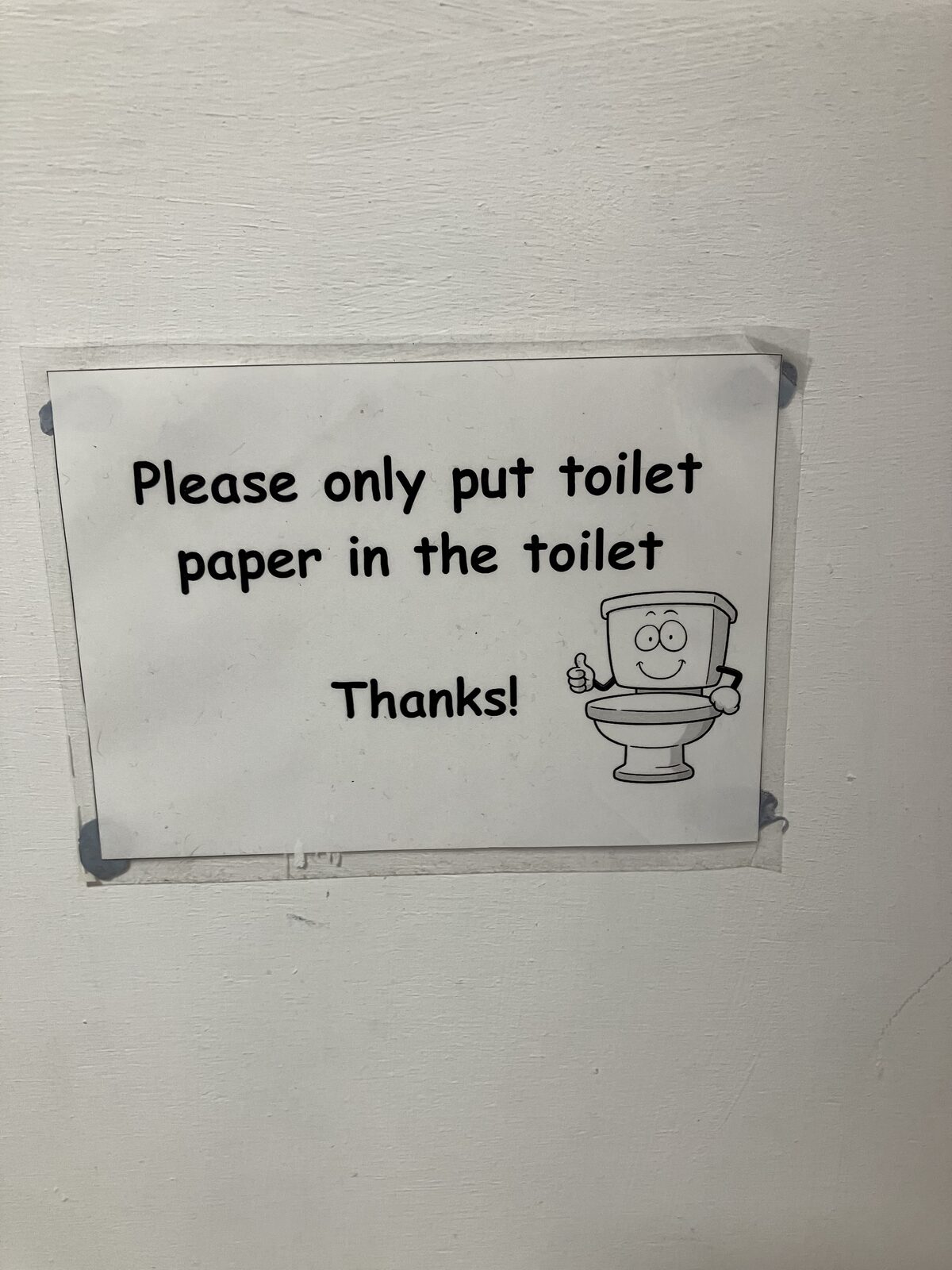 A simple white sign taped to a wall saying to put only toilet paper in the toilet, beside a smiling cartoon toilet giving a thumbs-up.