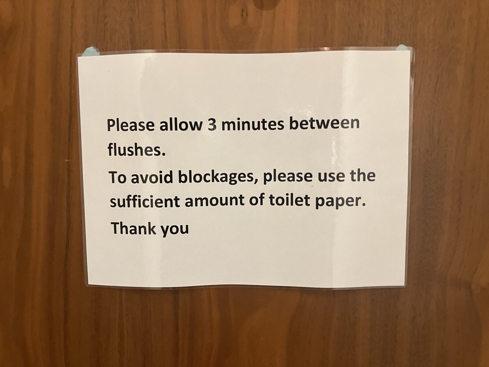 A laminated white sign on a wood door asking users to allow three minutes between flushes and to use a sufficient amount of toilet paper.