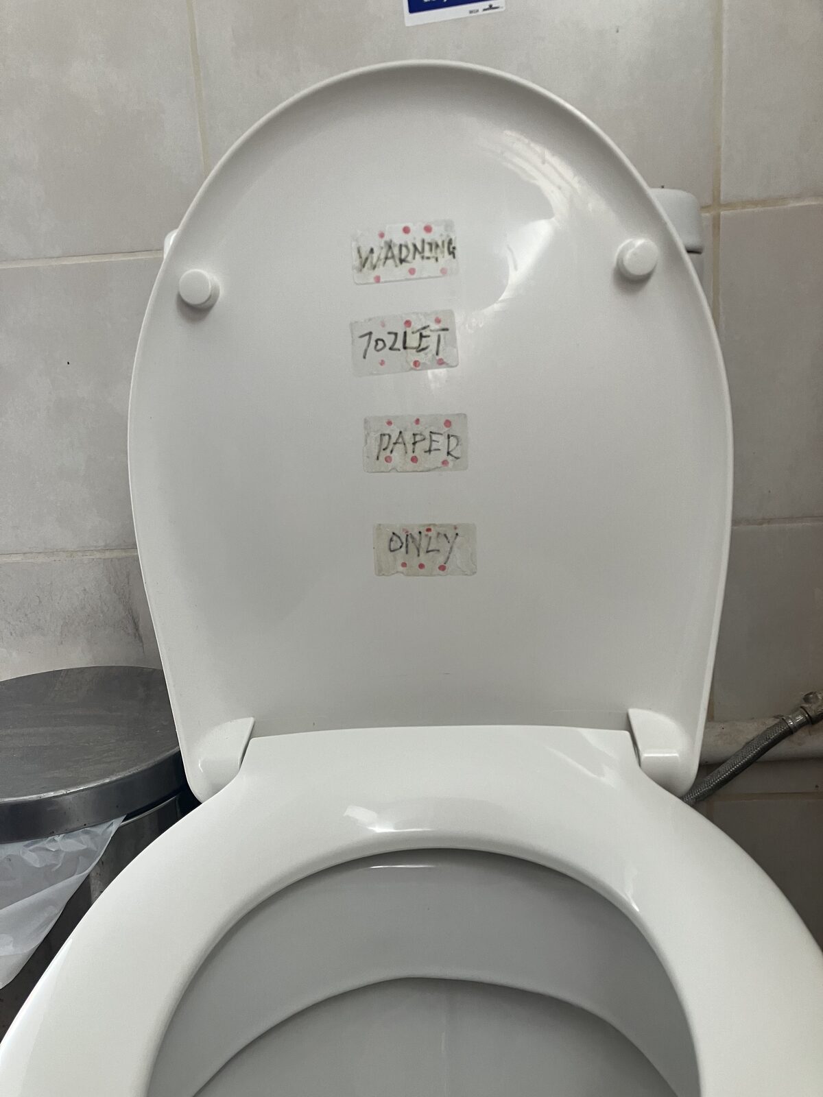 A white toilet seat lid with four small strips of handwritten tape reading 'WARNING', 'TOILET', 'PAPER', and 'ONLY'.