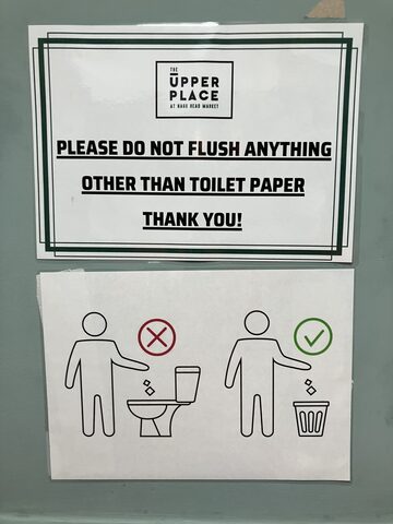 Two signs on a pale green wall: the upper sign says not to flush anything other than toilet paper, and the lower sign shows a pictogram placing waste in a bin rather than a toilet.