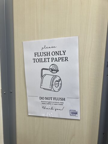 A printed sign on a light wood door with an illustration of a toilet roll, asking users to flush only toilet paper and not feminine hygiene products, baby wipes, rubbish, or paper towels.