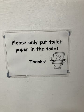 A simple white sign taped to a wall saying to put only toilet paper in the toilet, beside a smiling cartoon toilet giving a thumbs-up.