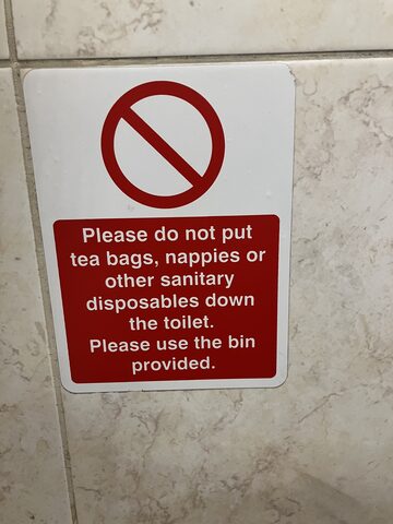 A red and white sign on a tiled restroom wall instructing visitors not to put tea bags, nappies, or other sanitary disposables down the toilet, and to use the bin instead.