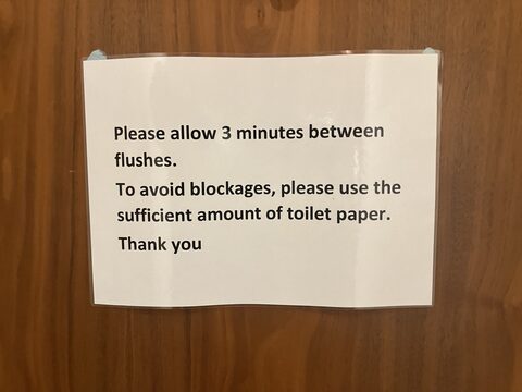 A laminated white sign on a wood door asking users to allow three minutes between flushes and to use a sufficient amount of toilet paper.