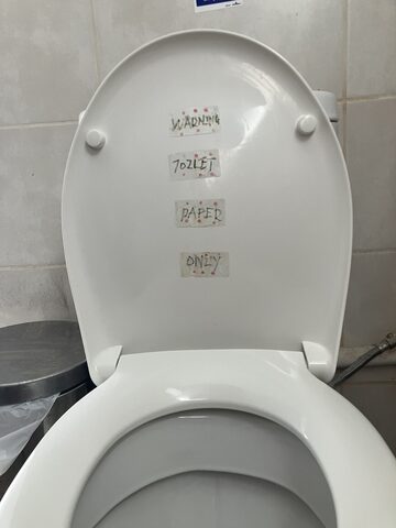 A white toilet seat lid with four small strips of handwritten tape reading 'WARNING', 'TOILET', 'PAPER', and 'ONLY'.